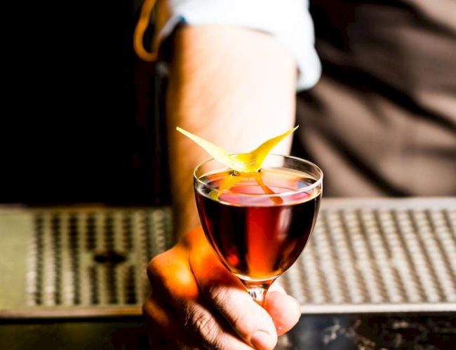 A person is holding a cocktail garnished with a yellow twist of citrus peel, standing behind a bar counter.